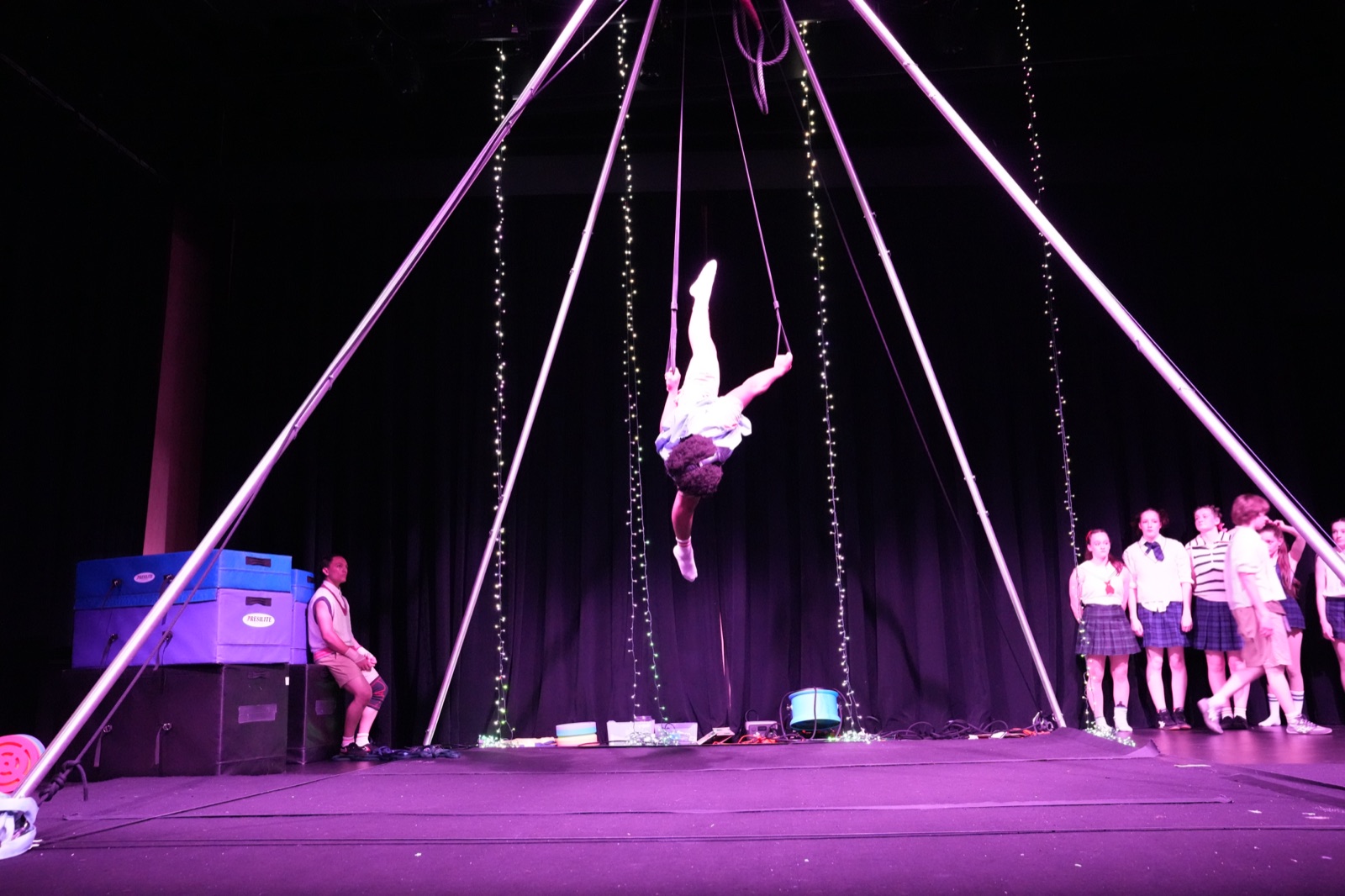 Aerialist inverted on the rig under purple light as the ALPHABET cast watches from the wings